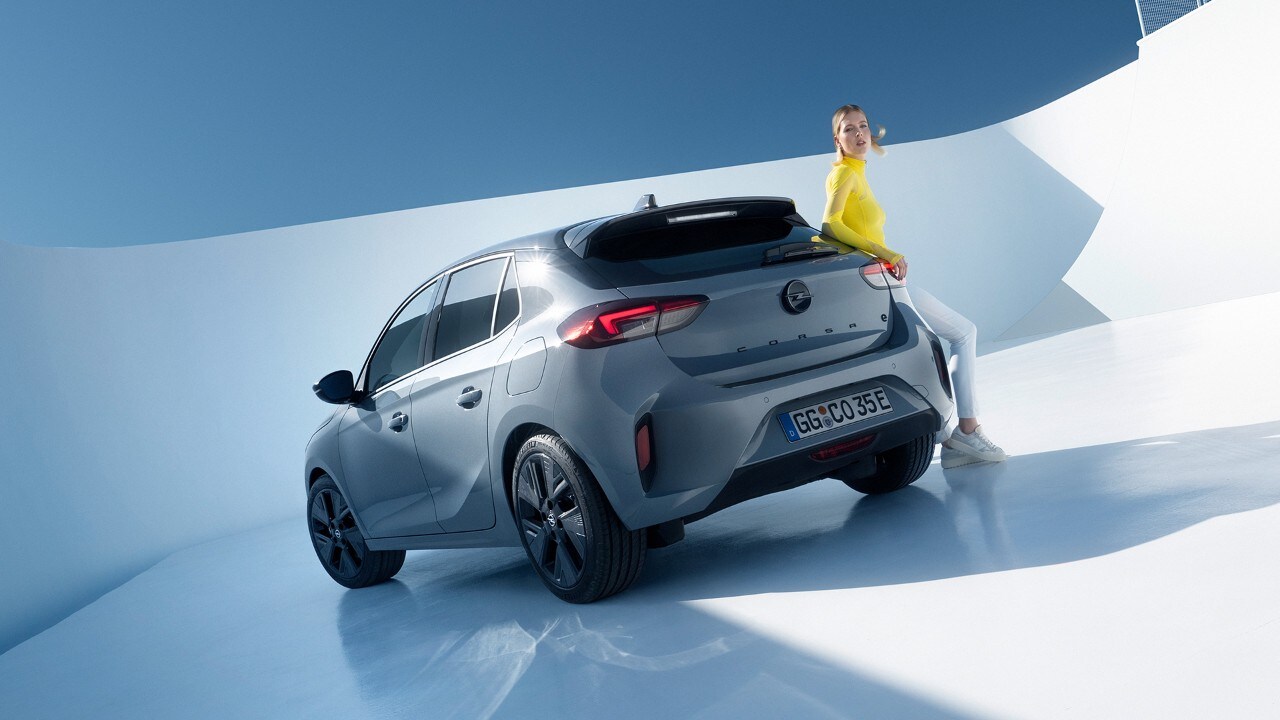 Vista lateral trasera de un Opel Corsa Electric gris con aleaciones negras y techo con un hombre apoyado contra él