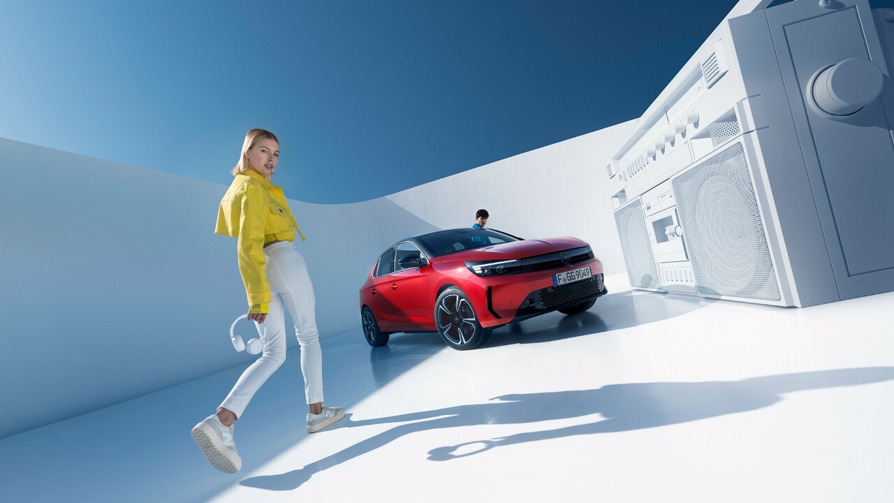 Opel New Corsa rojo aparcado en la distancia con mujeres con chaquetas amarillas caminando hacia él y mirando a la cámara